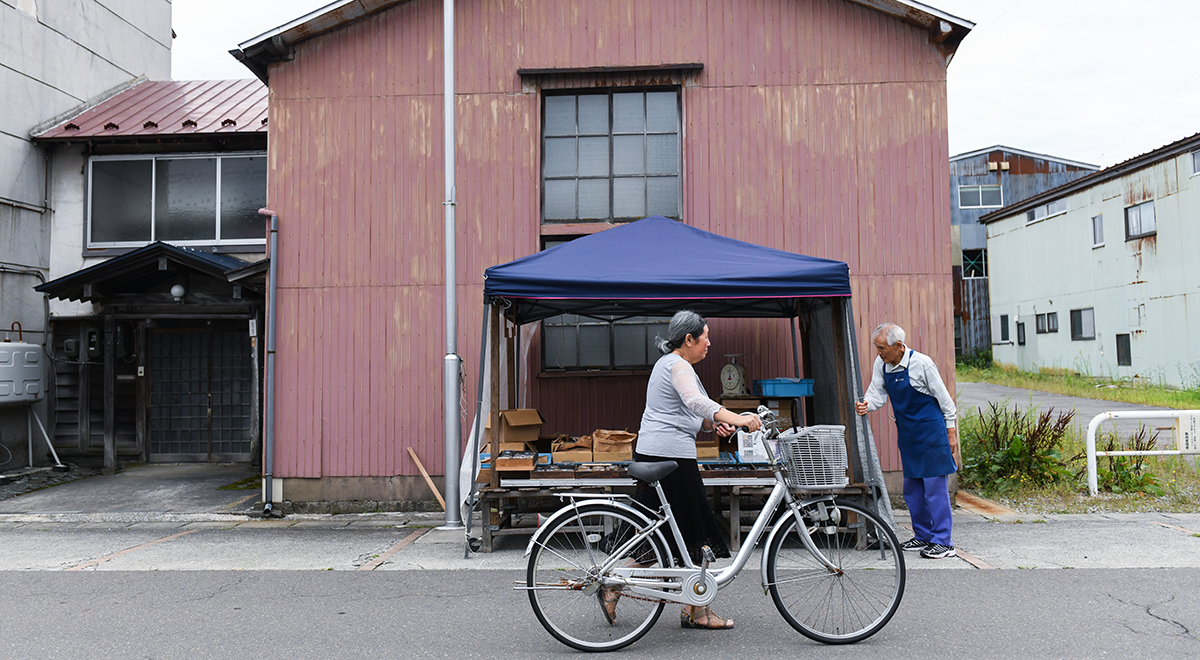 A woman pushes a bicycle past a vendor at a morning market in Gojome