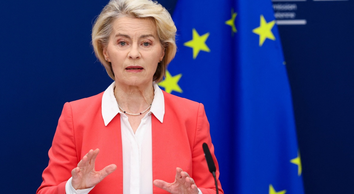 Ursula von der Leyen, pink suit, white collar shirt, blonde hair, black microphone, EU flag, stars