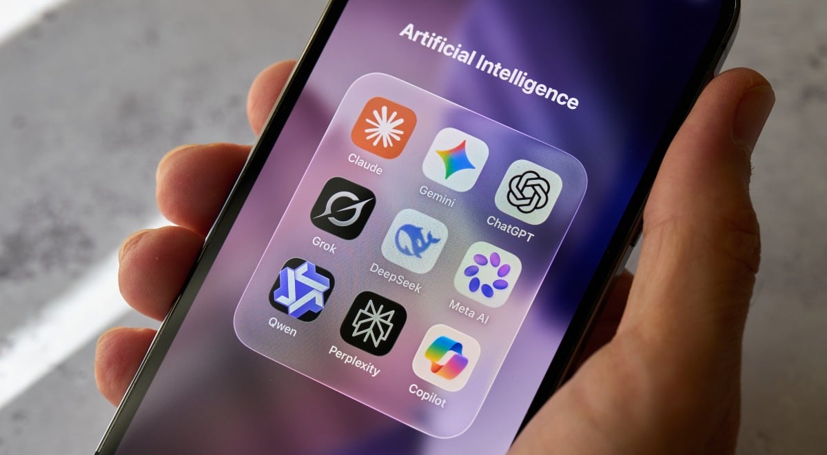 A hand holds a smartphone displaying the Claude, Gemini, ChatGPT and other AI apps