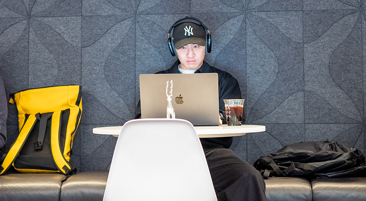 Man wearing headphones and baseball cap working on laptop at cafe table with backpack and iced coffee