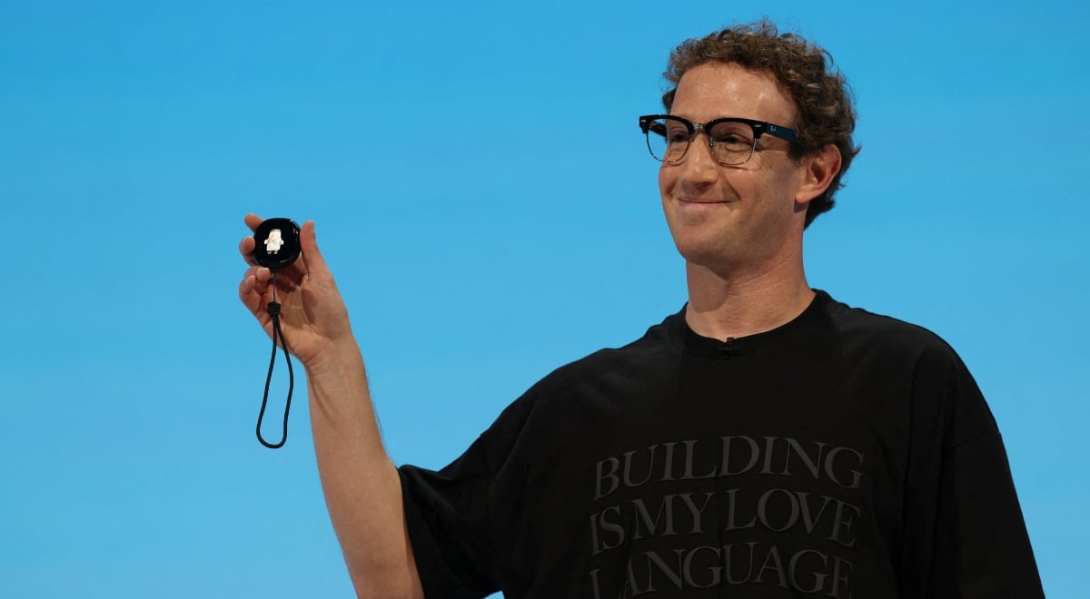 Zuckerberg, curly hair, black tshirt, baby blue background, small round gadget in raised right hand