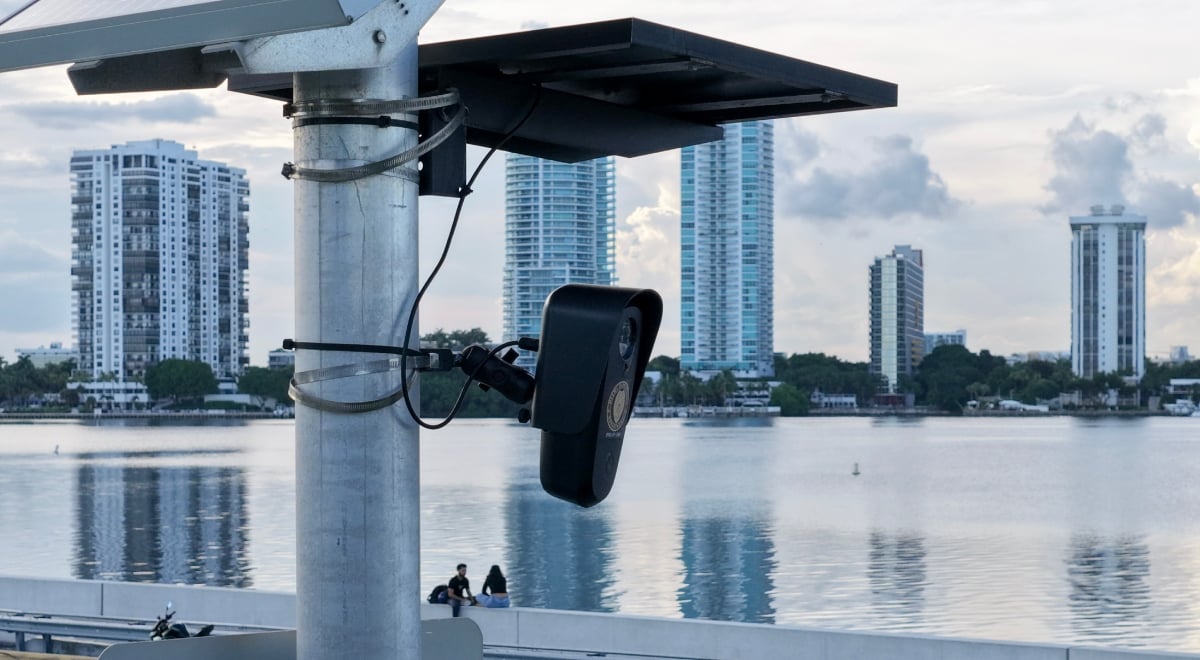 black flock camera, two solar panels above it, blue water, five skyscrapers, blue cloudy sky, couple