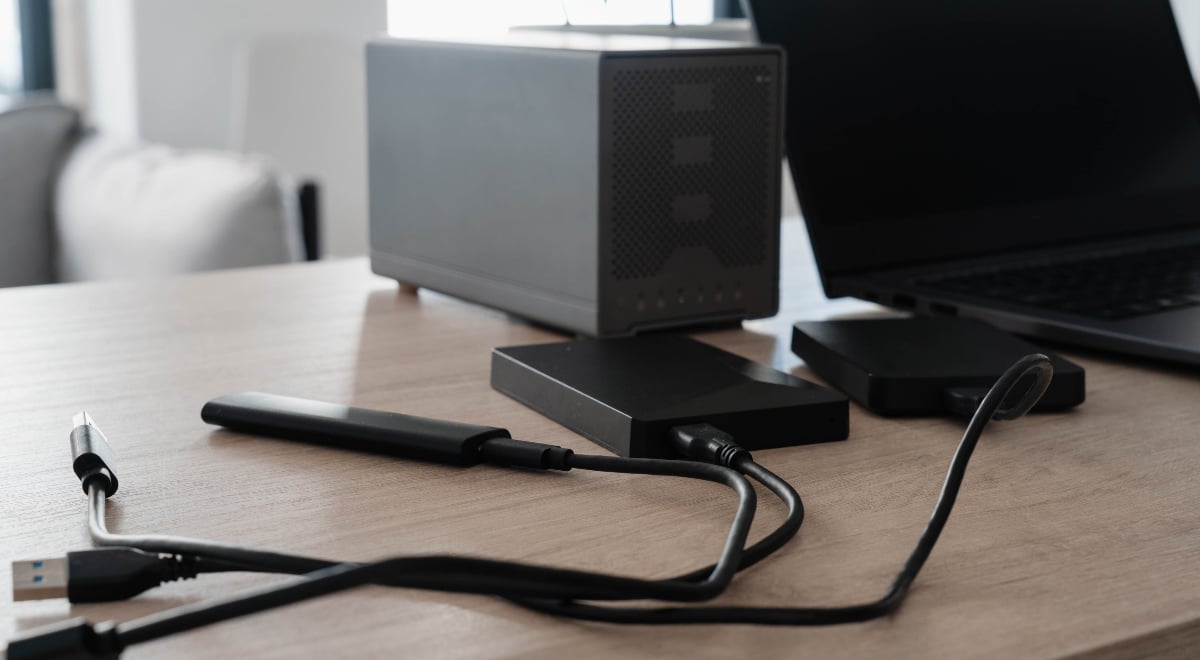 External hard drives, a portable SSD and USB cables connect to a laptop on a wooden desk
