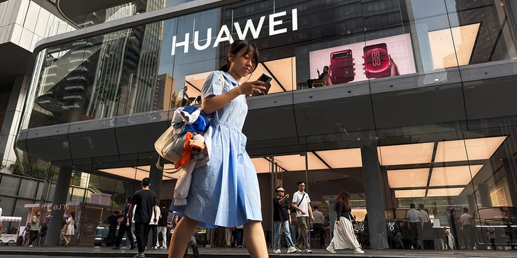 A pedestrian looks at a smartphone while walking past a Huawei retail store displaying an illuminated Huawei sign and smartphone advertisements 