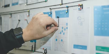 A man's hand pointing with a pin to a map hung on a wall