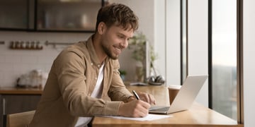 Close up smiling young man using laptop, writing, taking notes, watching webinar, training, involved