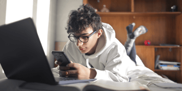 Teenage boy using mobile and laptop while lying on bed
