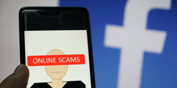 Mobile phone with scam written on it and FB logo in backdrop