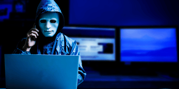 Man in sinister white mask sitting at computer on a mobile phone