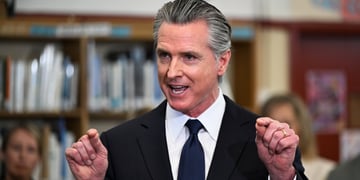 Gavin Newsom, black suit and tie, fists up, grey combed back hair, library, books, smiling, talking