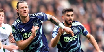 two football players run, dark blue tops, ajax logo, crowded stadium in the back, long blond hair