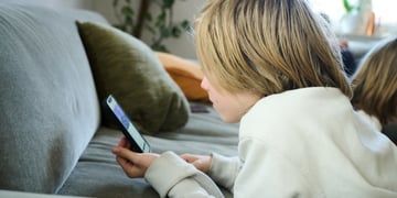 blonde boy, shoulder length hair, scrolls smartphone, white sweatshirt, grey sofa