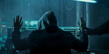 dark web hacker in hoodie, arms up, left arm handcuffed, arrest, dark blue light room