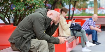 bald man, scrolls phone, red bench, dark green jacket, green trees