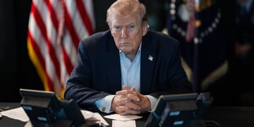 president donald trump sitsat desk, shadowy office, us flag in the back, blue shirt, no tis