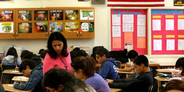 classroom, teacher in pink top, isa flag, kids leaning on tables, 