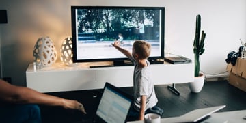 noy kneeling in front of smart tv, points to screen, living room, white wall, green cactus