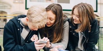 two teen girls, brunettes hold smartphones, one blonde teen boy, wait in bus stop, laugh