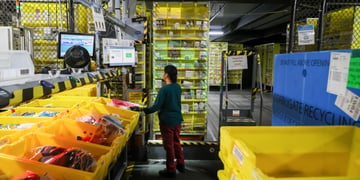 Female worker, dark hair, Amazon fulfillment center, yellow parcel boxes, fence, dark red throusers