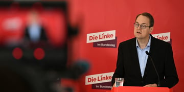 Soeren Pellmann, man in black suit, light blue shirt, no tie, collar unbuttoned, red photo wall