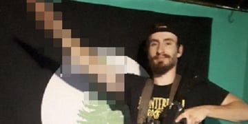 white man, black beard, moustache, salutes, black tshirt, yellow sign, brown belt, dark rifle, firn 