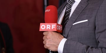 grey suit, white shirt, grey tie with stripes, dark orange microphone, ORF white letters, white hand