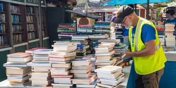 old man, live yellow reflective vest, blue tshirt, piles of books, shelves of books, tan, brown shorts