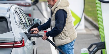silver electric car, black charging cable, smiling old man in jeans, black jumper, sand color vest