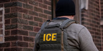 ice agent from back, black hat, white male, grey top, yellow letters ICE, dark brown brick wall 