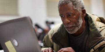 Black man in grey hair, moustache, and beard, grey laptop, army green winter jacket, 