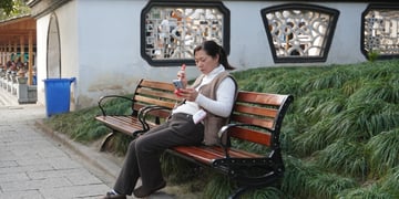 old chinese woman, black hair, wooden bench, smartphone, white building wall, green bushes 