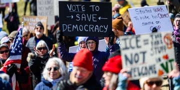 crowd of people holding protest signs, black sign, white letters, vote to save democracy
