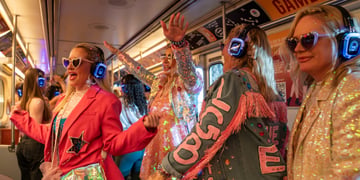 four blonde women wear blue whiny headphones, sparkly outfits, on a white bus, pink jacket