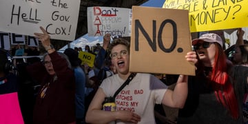 white woman, short curly blonde hair, protest signs, many people, yellow framed glasses