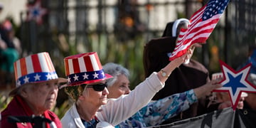 elderly white American couple, tall hat with US flag, black sunglasses, black fence, white jacket