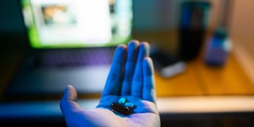 two white, two brown pills in white male hand, open laptop screen, brown desk.