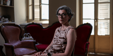 italian woman, sleevless shirt, grey hair, black frame glasses, tall windows, vintage sofa