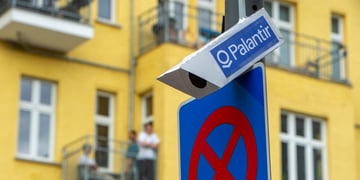 white surveillance camera on grey poll, blue sticker with palantir written on it, yellow building