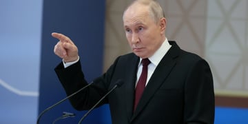 putin points right hand finger, black suit, white collar shirt, bordeaux tie, blue wall