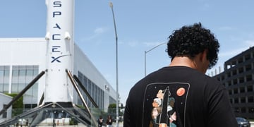 spaceX white rocket, white building, black man, black t-shirt with rocket, black curly hair, blue sky 