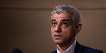 Sadiq Khan looking up, grey hair, black suit, white collar shirt, dark brown background, grey hair