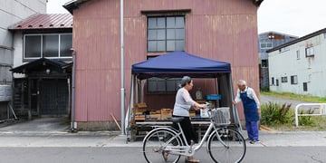 A woman pushes a bicycle past a vendor at a morning market in Gojome