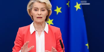 Ursula von der Leyen, pink suit, white collar shirt, blonde hair, black microphone, EU flag, stars