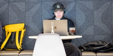 Man wearing headphones and baseball cap working on laptop at cafe table with backpack and iced coffee
