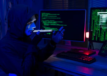 hacker, black hoody, white plastic mask on face, smartphone, two screens with green numbers, code