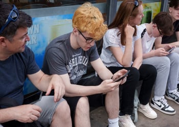 yellow curly hair, young white teen, grey tshirt, smartphone, three other people, glasses