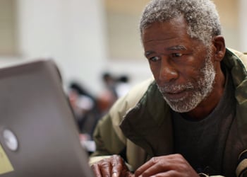 Black man in grey hair, moustache, and beard, grey laptop, army green winter jacket, 