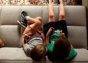 two white boys, green t-shirt- black shorts, grey t-shirt, light grey sofa, roblox on iphone screen