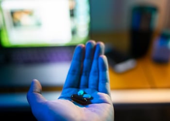 two white, two brown pills in white male hand, open laptop screen, brown desk.