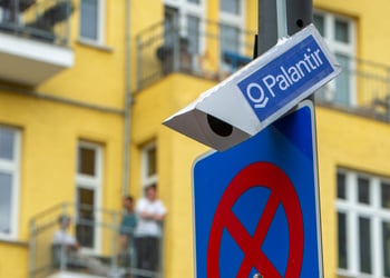 white surveillance camera on grey poll, blue sticker with palantir written on it, yellow building